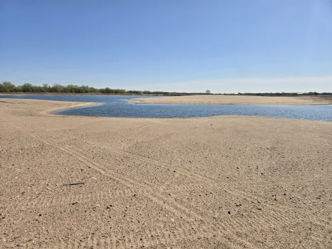 Wide view of a sandy shoreline and shallow blue water at the Newark West Sandpit in Nebraska, with a tree line in the distance under a clear sky.