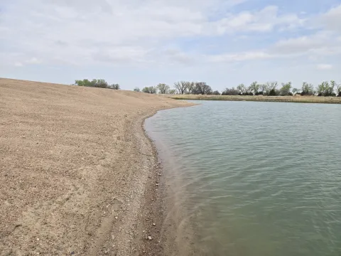 View of a gently sloped sandy shoreline curving around a calm off-channel basin, with trees and cloudy sky in the background at Cottonwood Ranch in Nebraska.