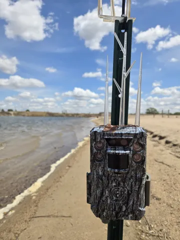 Close-up of a camouflaged trail camera attached to a metal post, with sandy shoreline and water in the background under a bright, partly cloudy sky.