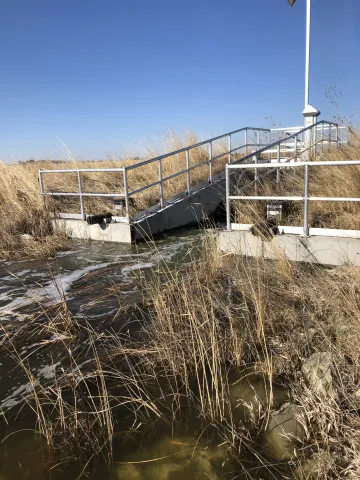 Water flowing beneath a metal access platform and hydraulic control structure beside tall grasses.