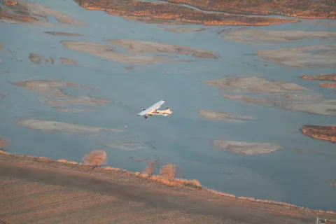 Small aircraft flying above braided river channels during early morning light.