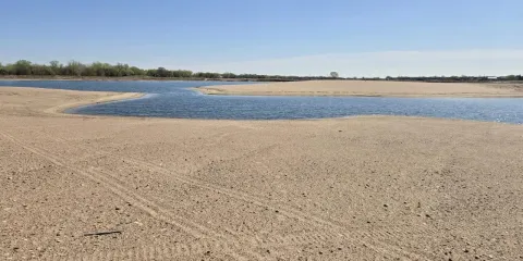 Wide view of a sandy shoreline and shallow blue water at the Newark West Sandpit in Nebraska, with a tree line in the distance under a clear sky.
